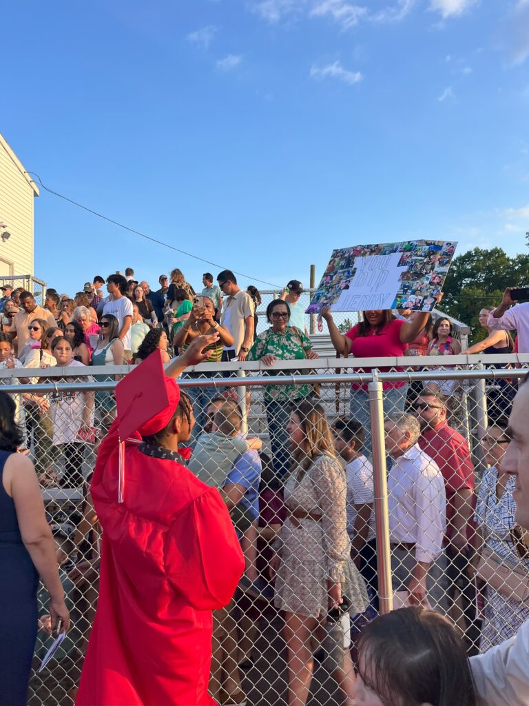 A photo of family members holding up a sign that reads "You did it, Jerry" on the bleachers while a graduate waves up to them.